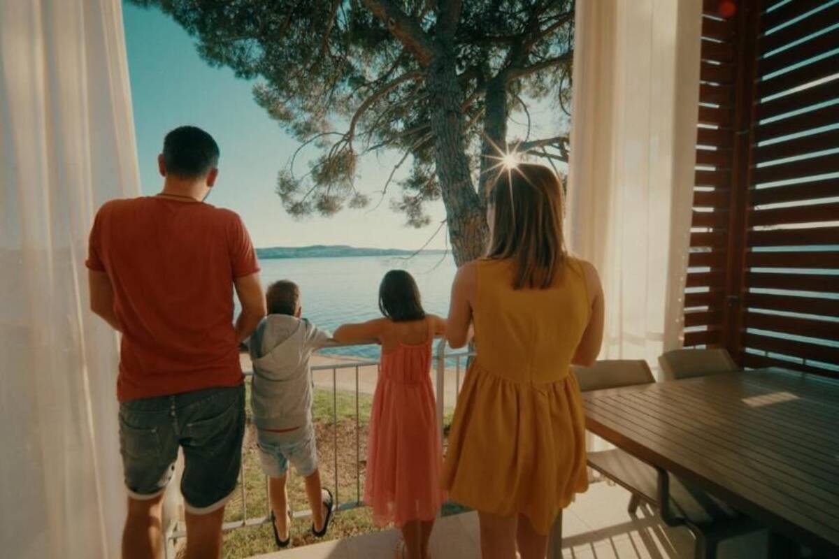 A family of four on a balcony overlooking a body of water, with a view of a tree and a distant shoreline.