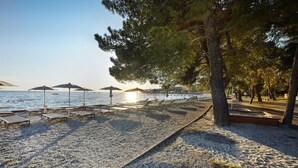 A beach with sun loungers and umbrellas, a pebble beach, and a row of trees.