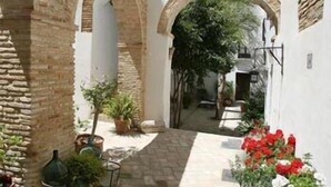 Un estrecho callejón con paredes blancas y arcos de piedra, que conduce a una zona cubierta con plantas en macetas y flores rojas.