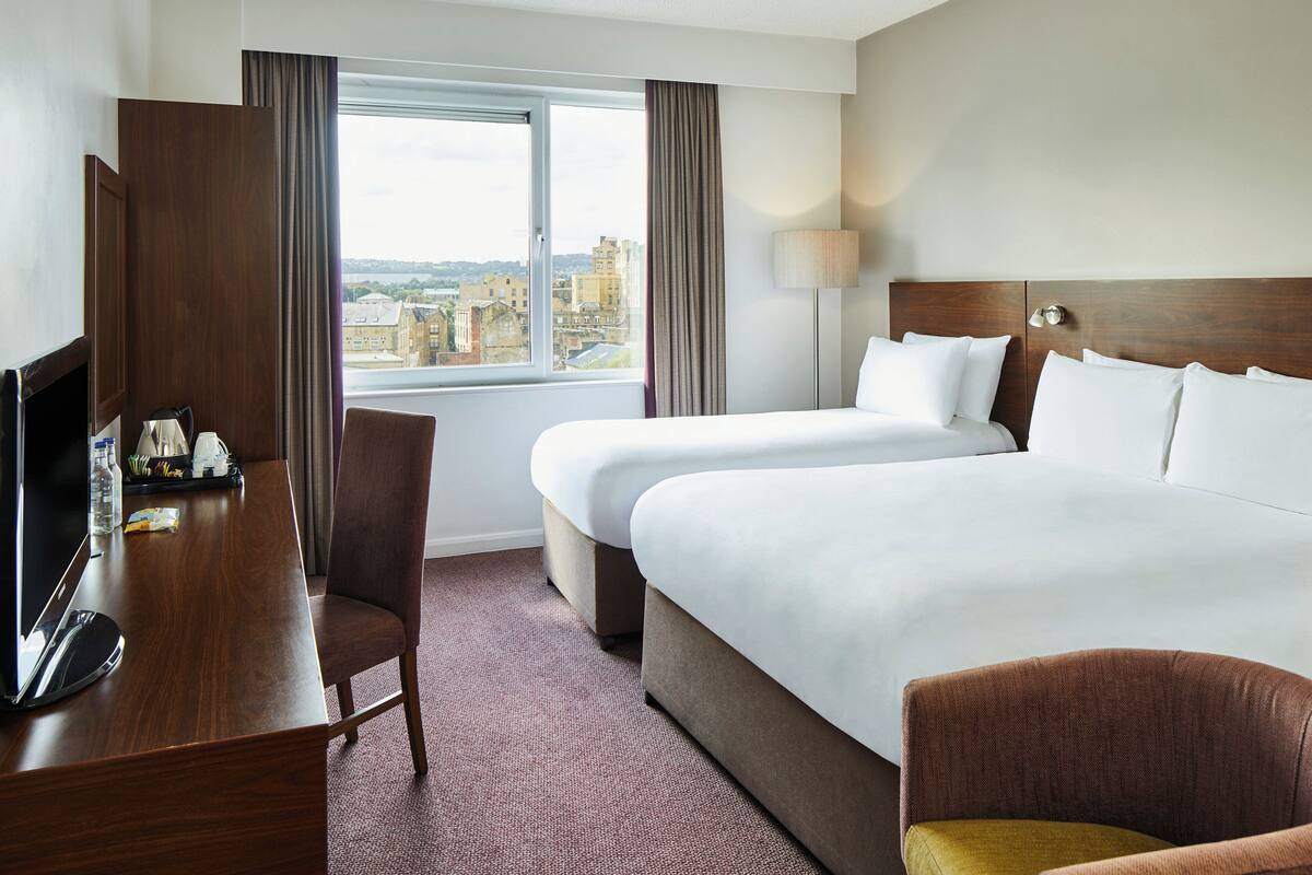 A hotel room with two beds, a desk with a TV, a chair, and a view of the city.