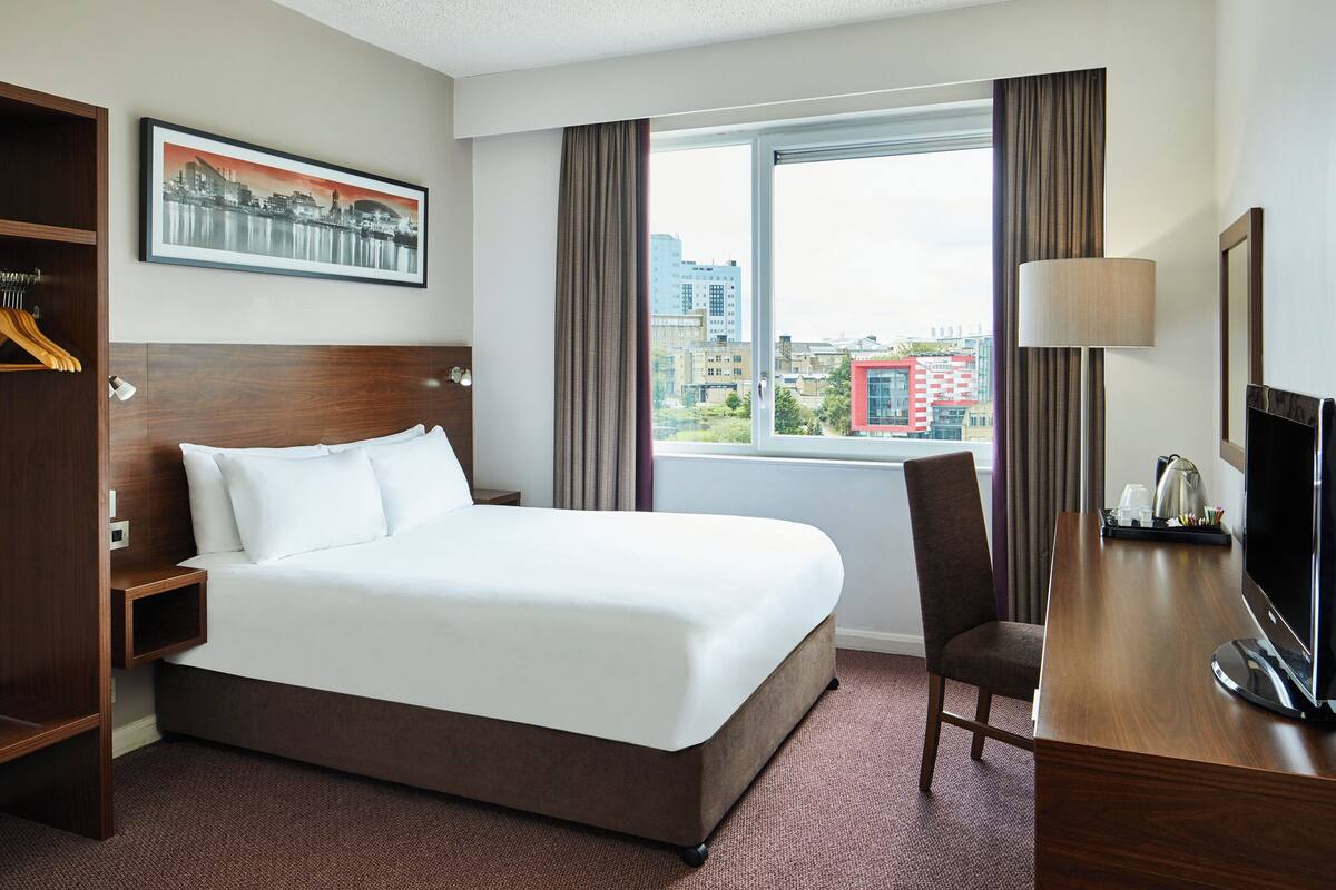A hotel room with a large bed, a desk with a television, a chair, a window with curtains, and a framed picture on the wall.