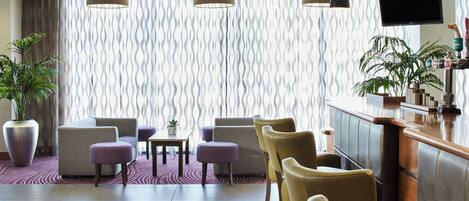 A modern hotel lobby with seating arrangements, a bar area, and a large window with patterned curtains.