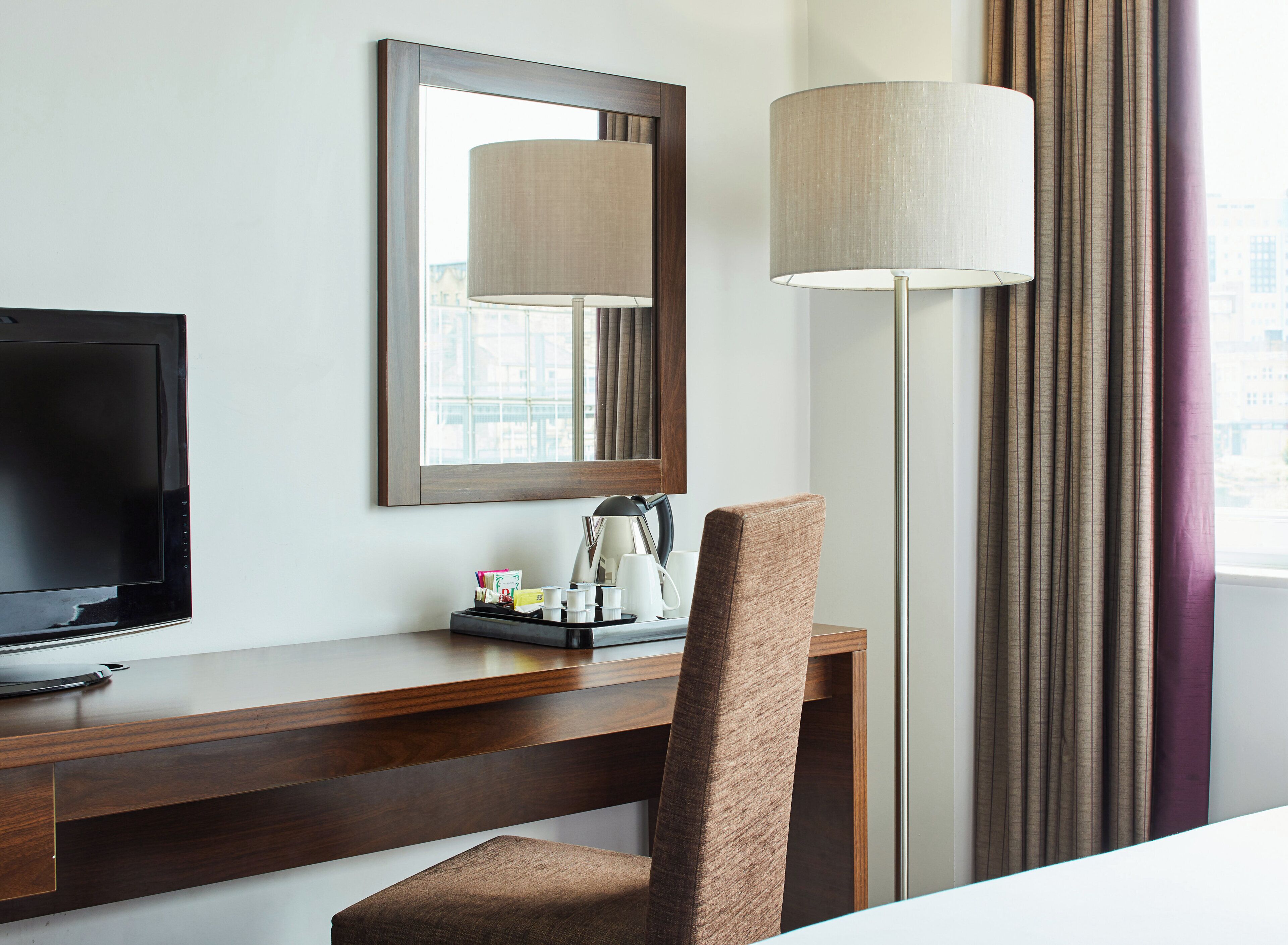 Une chambre d’hôtel avec un bureau en bois, une télévision, un miroir, une lampe et une chaise.