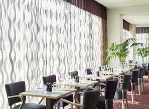 Un restaurante con una decoración moderna, que presenta separaciones blancas onduladas, sillas tapizadas en morado y mesas ordenadas con copas de vino y cubiertos.