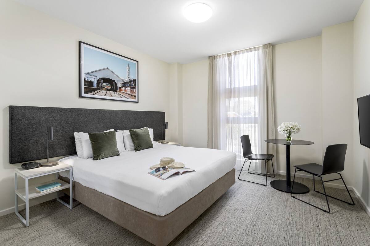 Habitación de hotel con una cama grande, mesita de noche, lámpara y una pequeña mesa con sillas.