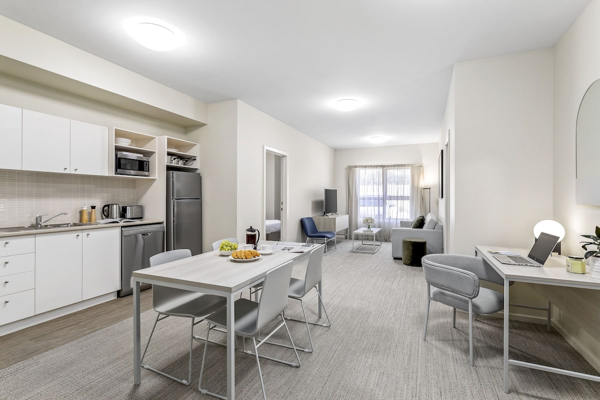 Una cocina moderna con armarios blancos, un refrigerador de acero inoxidable y un microondas. Hay un comedor con una mesa y sillas, y un pequeño espacio de trabajo con una computadora portátil y una lámpara.