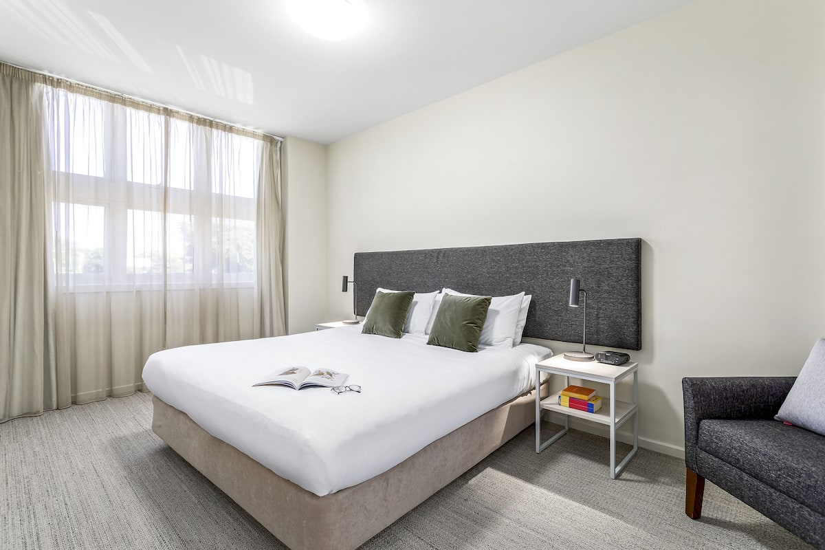 Habitación de hotel impecable con una cama grande, una mesita de noche con lámpara, una silla y vistas al exterior a través de cortinas translúcidas.