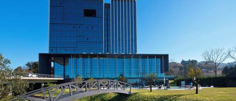 A modern hotel with a glass facade, a bridge, and a grassy area with outdoor seating.