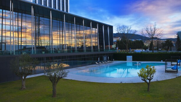 Un bâtiment moderne doté d’une grande façade en verre, d’une piscine et d’un espace paysager agrémenté d’arbres et de chaises longues.
