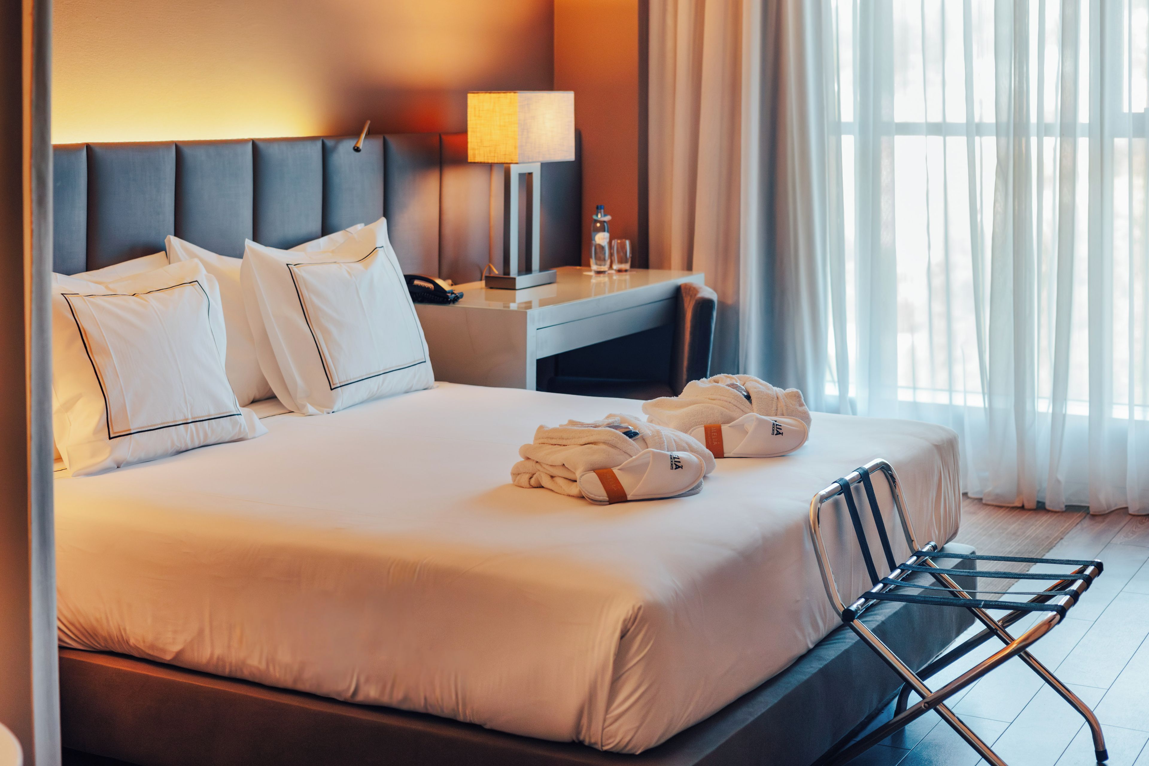 A hotel room with a large bed, a desk with a lamp, a chair, and a window with curtains.