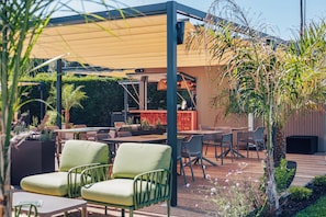 Un espacio al aire libre con mesas y sillas, una sección cubierta con una mesa y sillas, y un fondo de vegetación y un edificio.