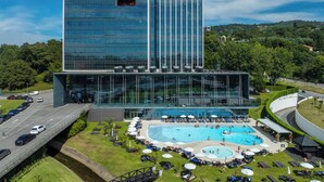 Un bâtiment moderne doté d’une façade en verre, d’une piscine sur le toit et d’un espace aménagé avec des chaises longues et des parasols.
