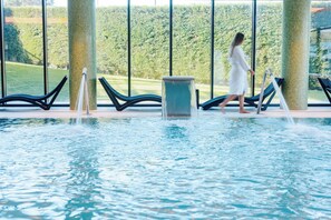 Un espace piscine où une personne nettoie l’eau, avec des chaises longues et une vue sur un paysage verdoyant à travers de grandes fenêtres.