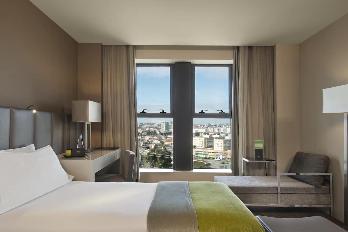 Een hotelkamer met een groot bed, een bureau, een stoel en uitzicht op de stad door grote ramen.