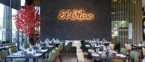 A restaurant interior with a brick wall, tables set for dining, and a neon sign reading 'El Olivo'.