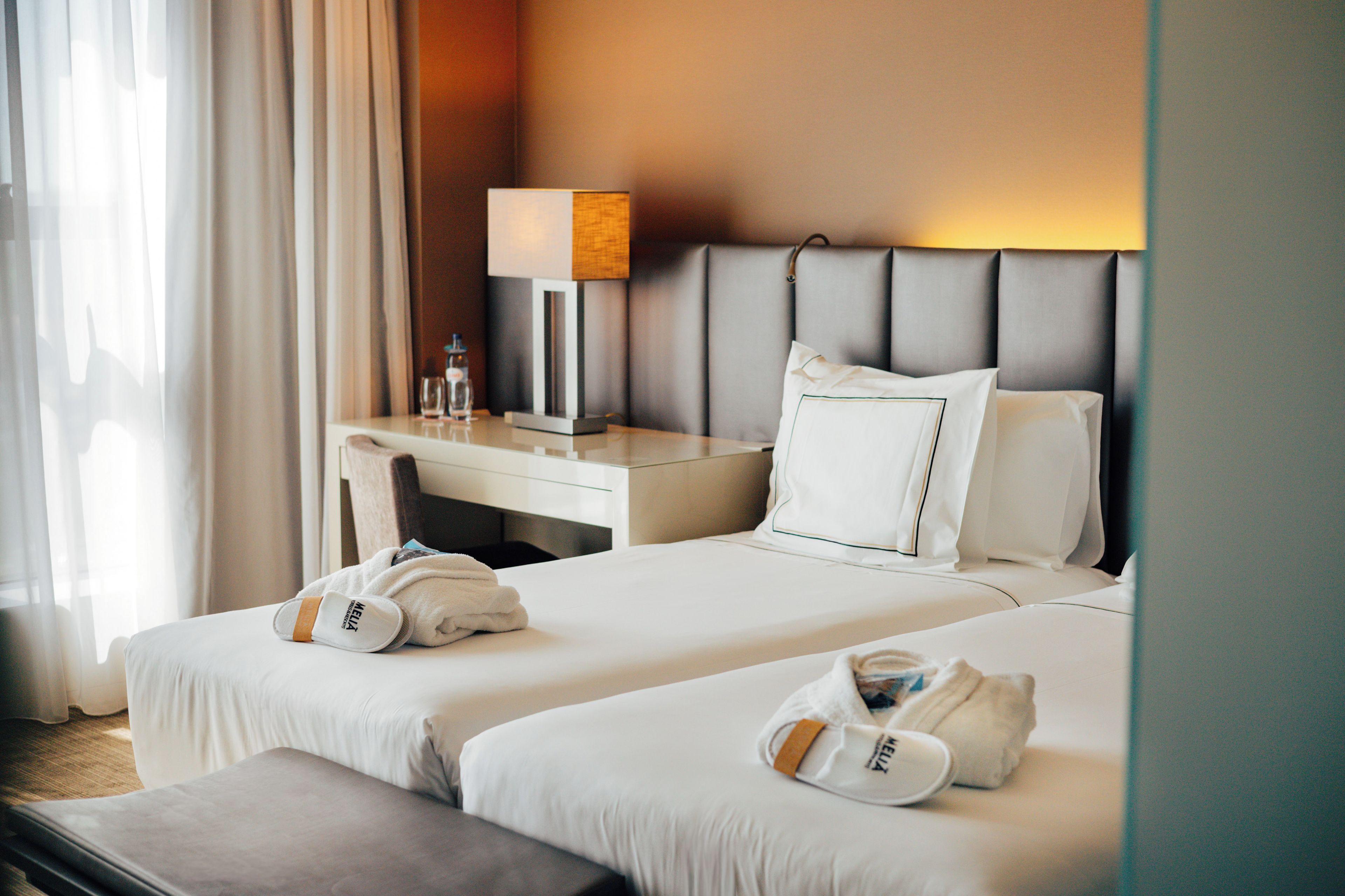 A hotel room with two beds, a desk, a lamp, and a window with curtains.