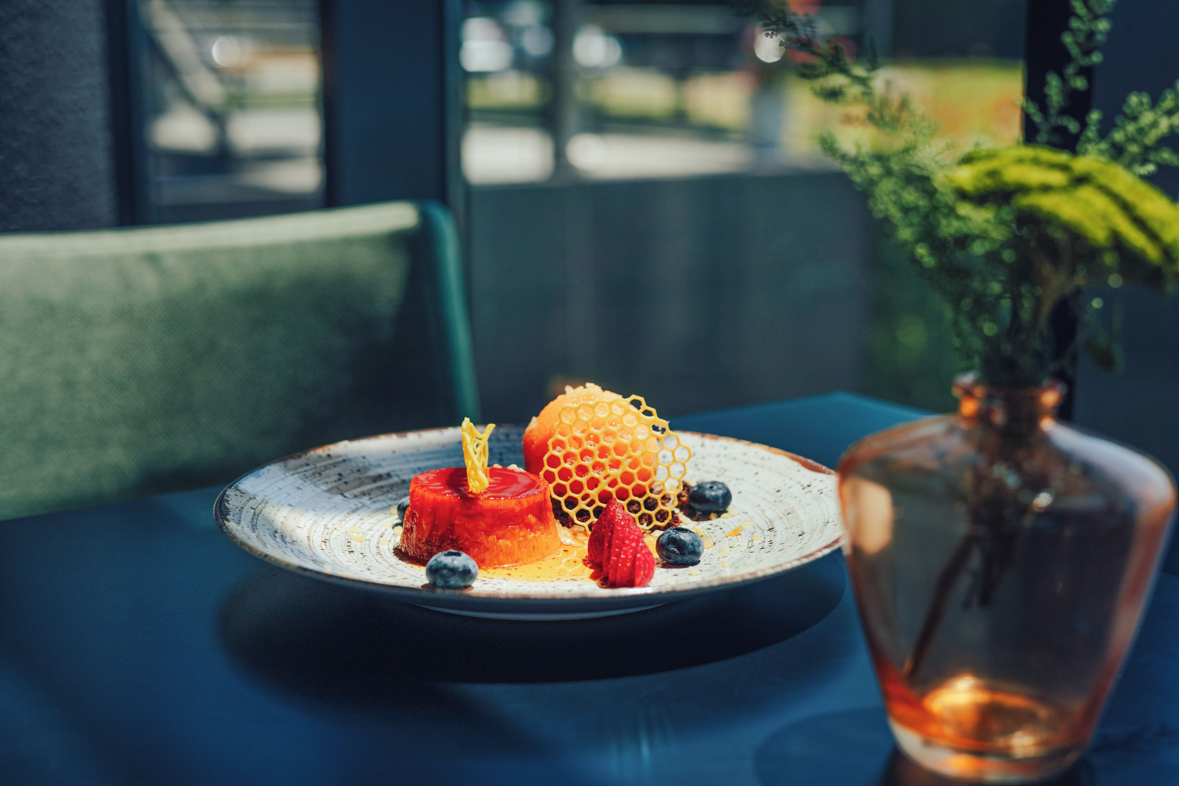 Un plato de postre con flan de caramelo, panal, fresas y arándanos sobre una mesa con mantel azul.