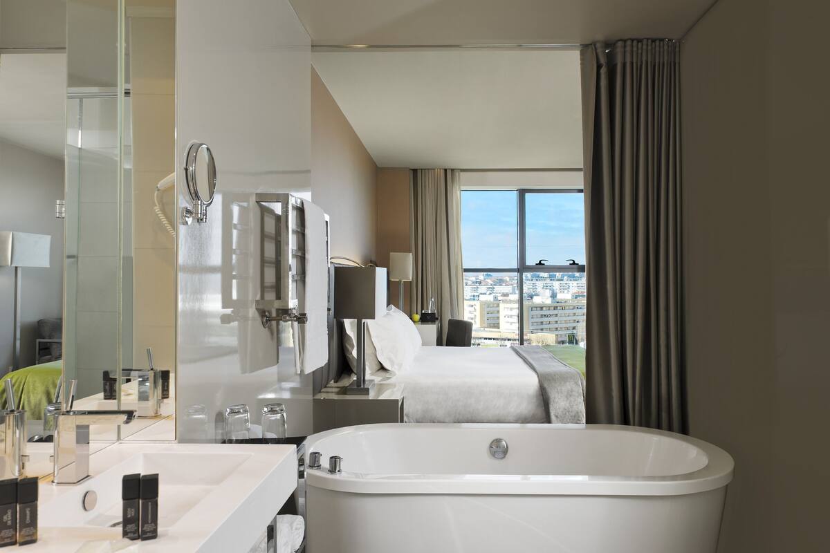 Een moderne hotelkamer met een groot bad, een aparte badkamer met spiegel en wastafel, een bed met uitzicht op de stad en een groot raam met gordijnen.