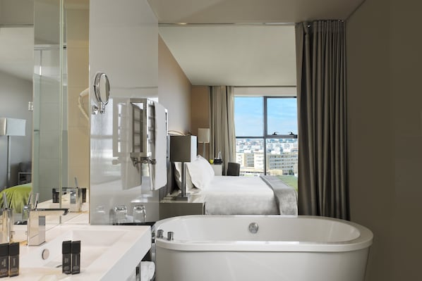 Une chambre d’hôtel moderne dotée d’une grande baignoire, d’une salle de bain séparée avec un miroir et un lavabo, d’un lit offrant une vue sur la ville et d’une grande fenêtre avec des rideaux.