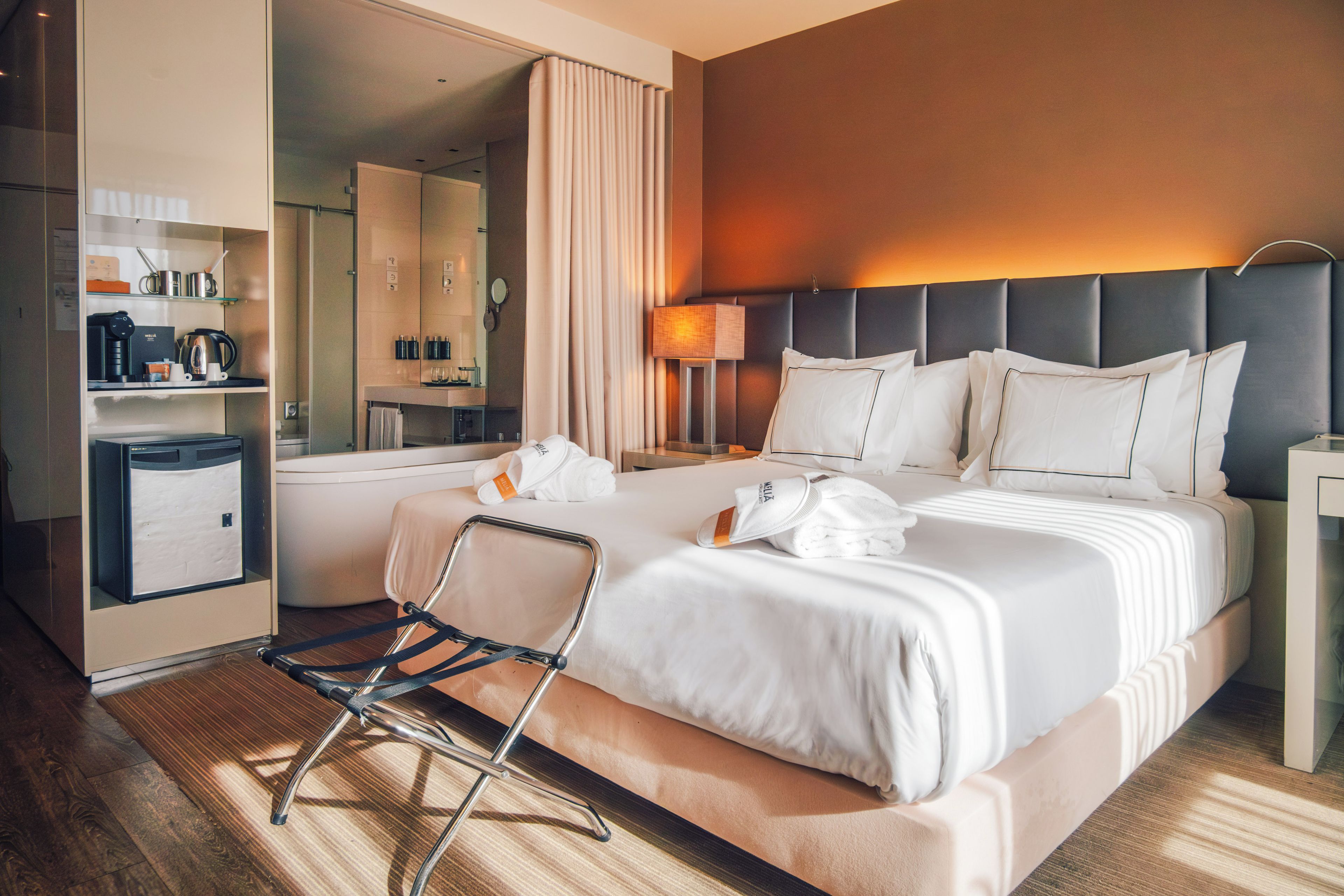 A hotel room with a bed, bedside tables, a chair, a desk with a lamp, a mirror, and a minibar.