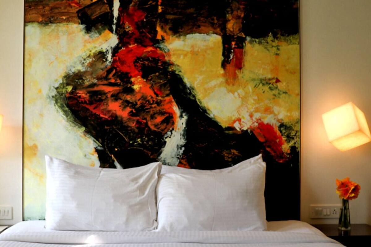 Una habitación de hotel con un cuadro abstracto grande en la pared, una cama con ropa blanca y una mesita de noche con una lámpara y un florero.