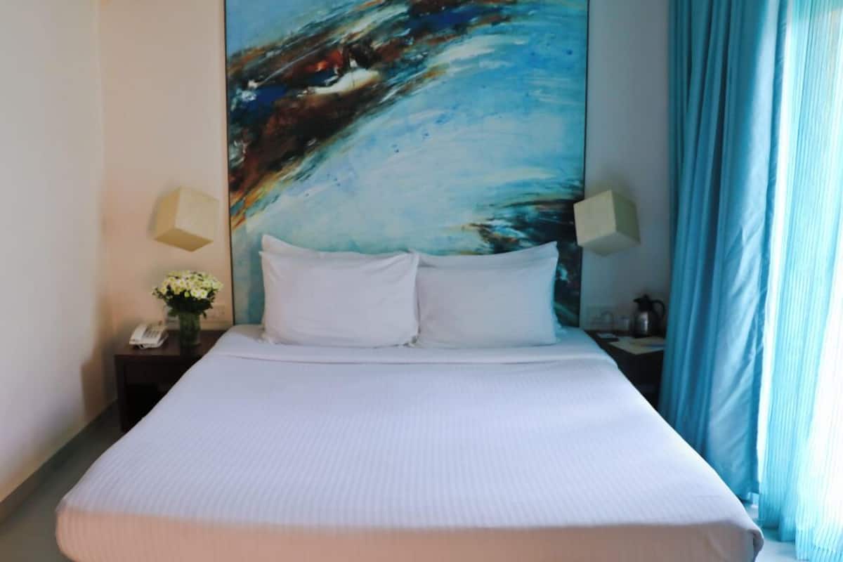 Una habitación de hotel con una cama grande, dos lámparas de noche, una mesita de noche con un teléfono y un jarrón de flores, y un cuadro en la pared.