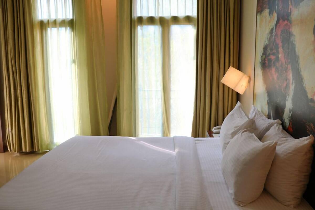 Habitación de hotel con una cama grande, almohadillas beige, una mesita de noche con lámpara y un cuadro en la pared.