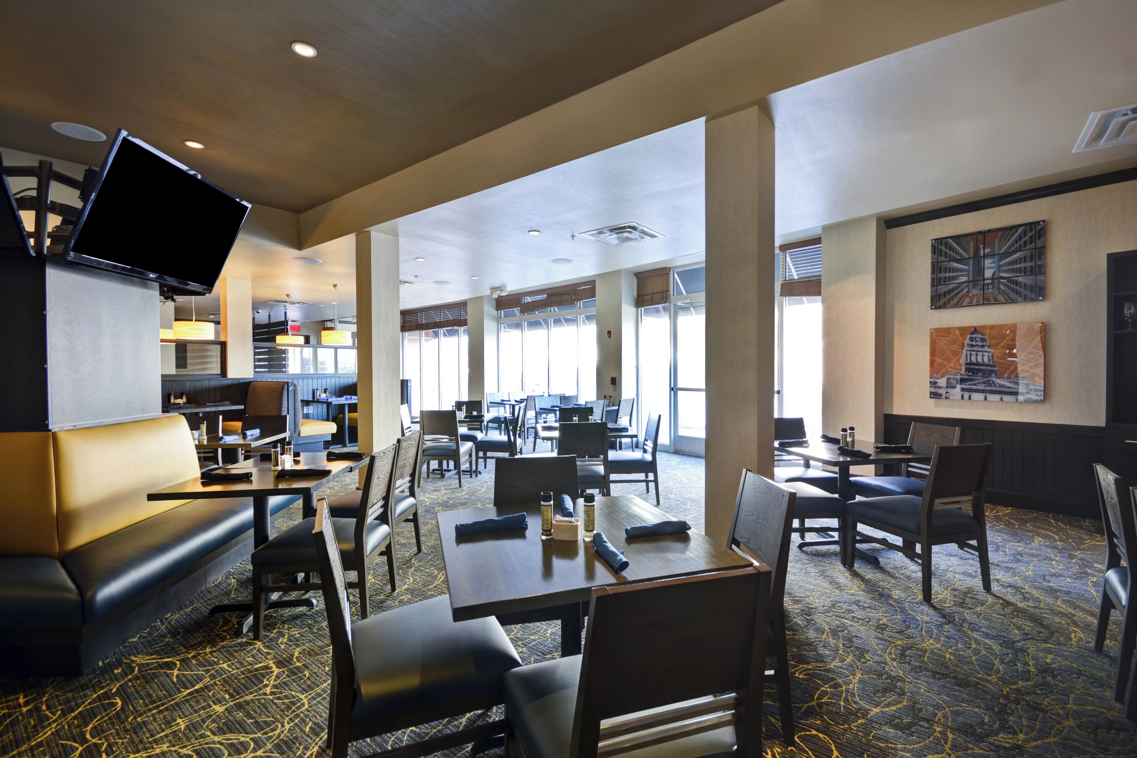 A hotel lobby with a television mounted on the wall, multiple seating areas including a booth and tables, and a large window providing natural light.