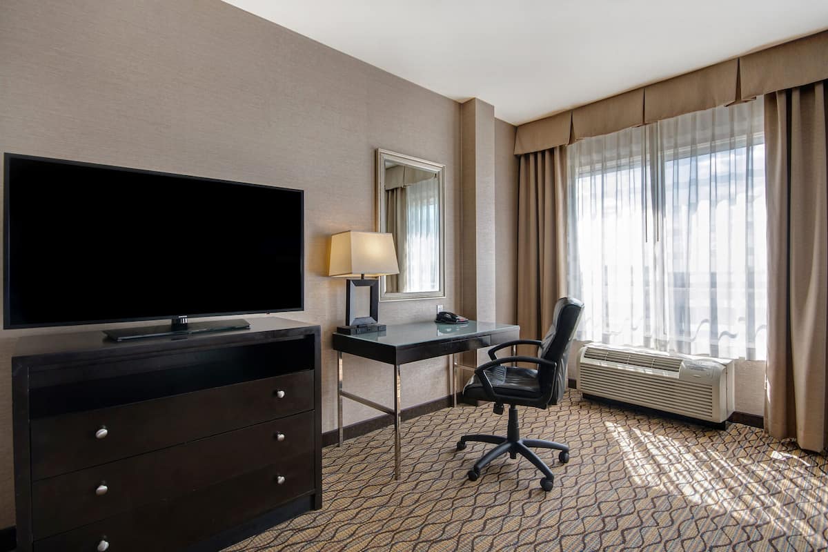 A hotel room with a flat-screen TV, a desk with a lamp, a chair, an air conditioner, and a large window with curtains.