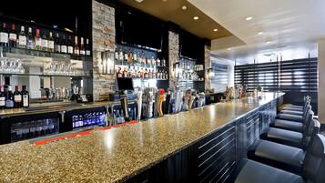 Un bar con encimera de granito, estantes con diferentes botellas de licor y una zona de estar con sillas negras.