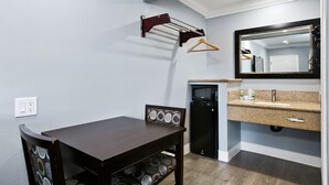 Ein kleines Hotelzimmer mit einem dunklen Holztisch, einem Stuhl mit gemustertem Rücken, einem kleinen schwarzen Kühlschrank, einer Arbeitsplatte mit Spüle und einem Spiegel.
