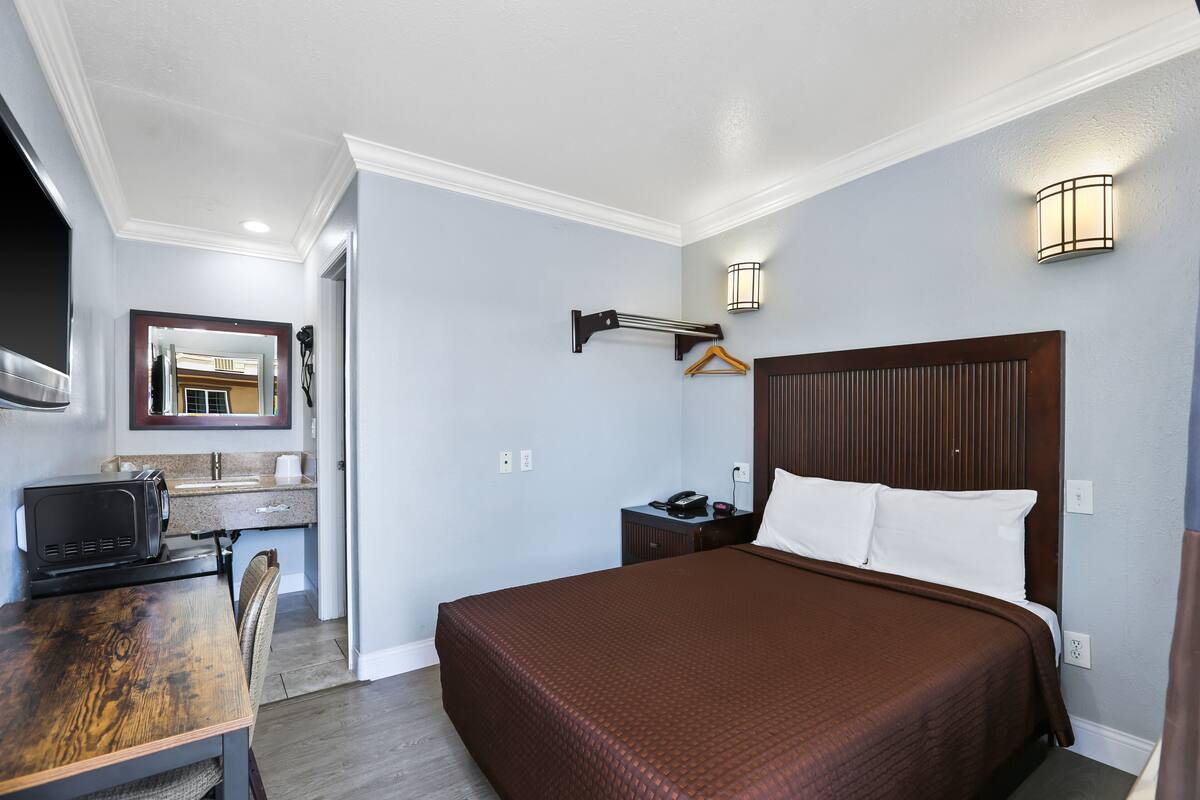 A hotel room with a bed, a desk with a microwave, a hanging coat rack, a bedside table with a lamp, and a mirror on the wall.