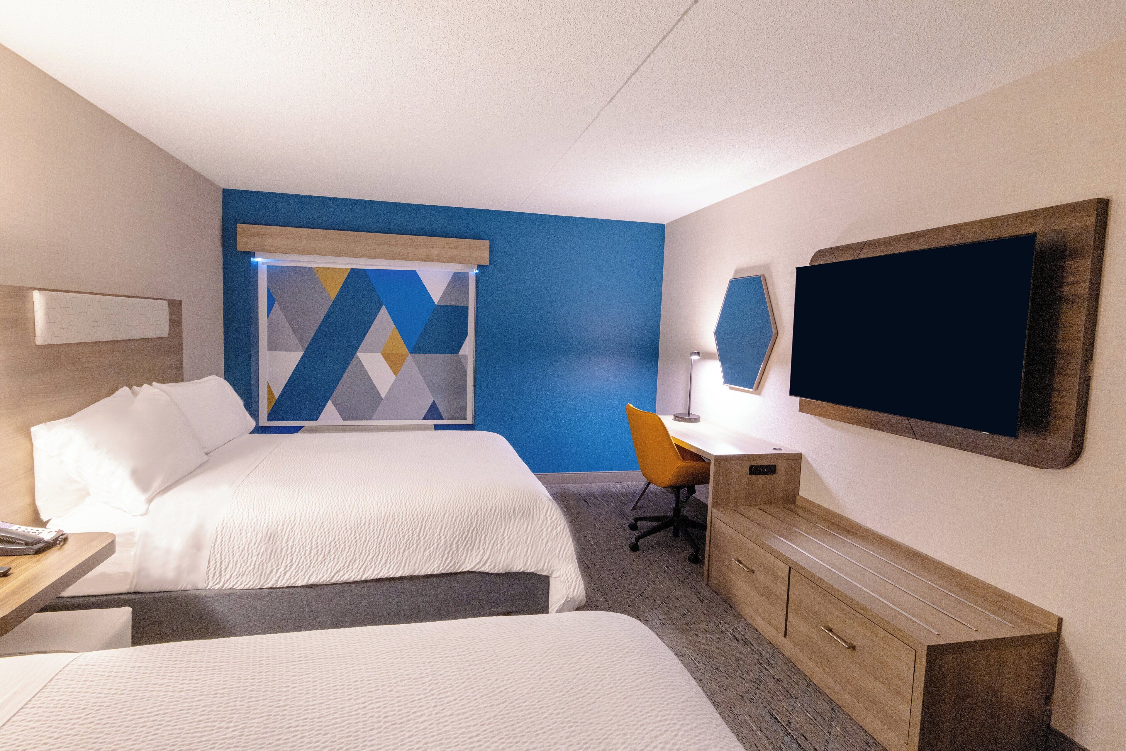 A hotel room with two beds, a desk, a chair, a TV, and a window with a geometric curtain.