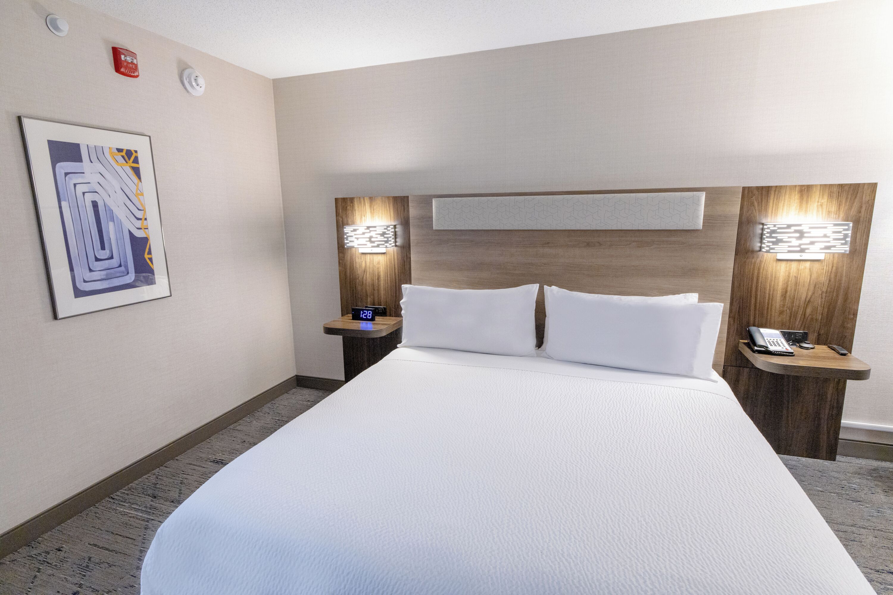 A hotel room with a large bed, two bedside tables with lamps, a clock, and a framed abstract painting on the wall.