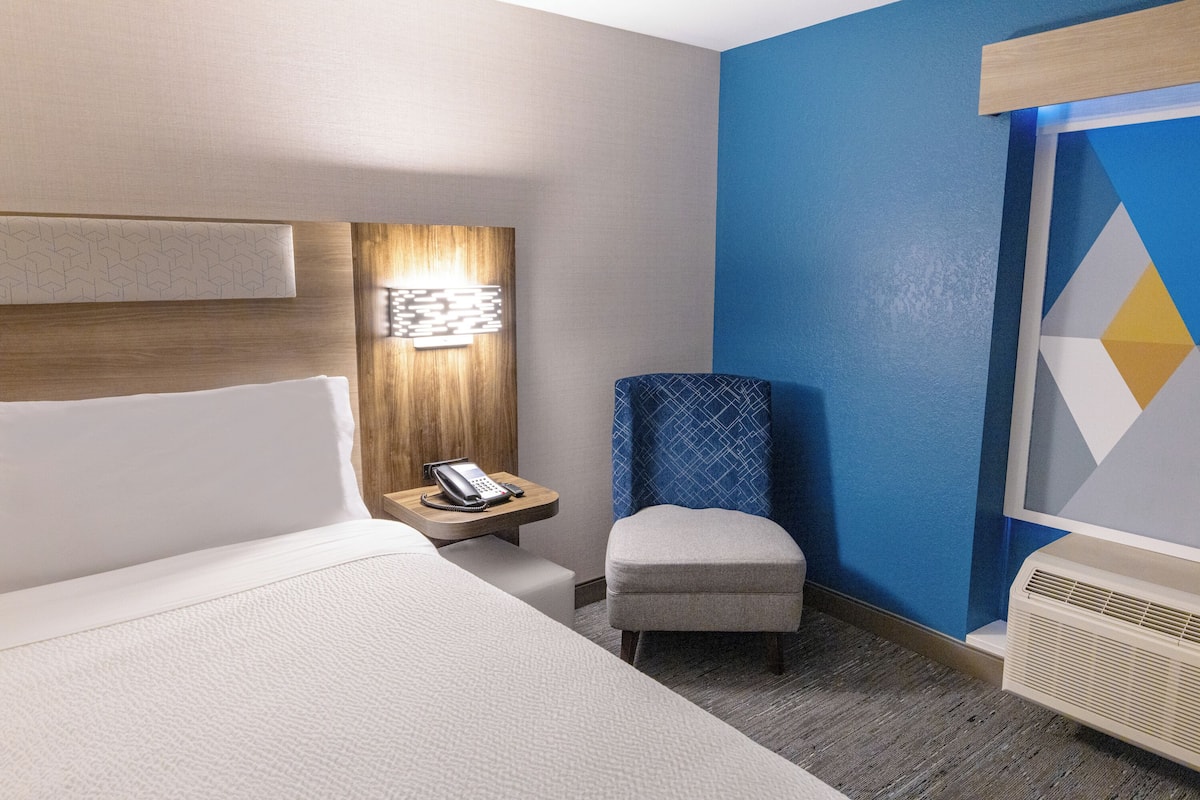 A hotel room with a bed, a small table with a phone, a chair, and a wall-mounted light fixture.