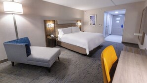 A hotel room with a bed, a chair, a table, a nightstand, and a lamp.