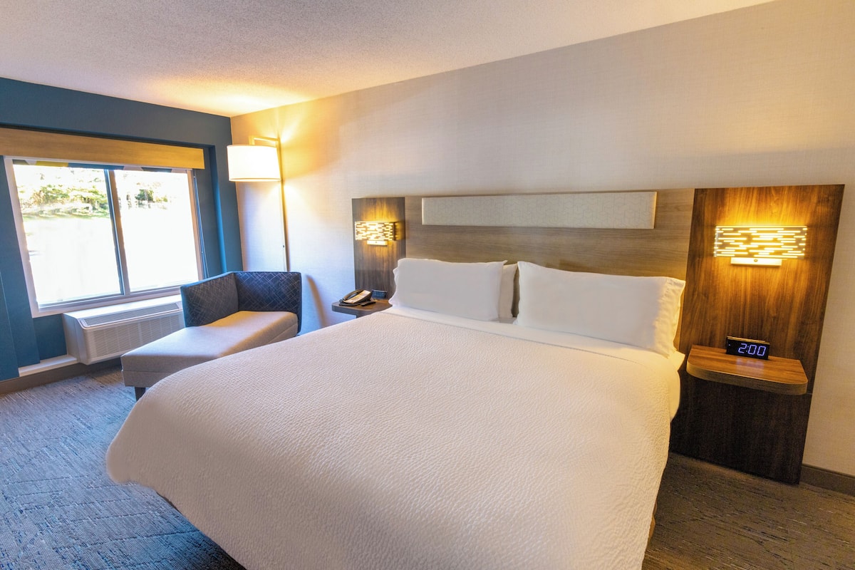A hotel room with a large bed, a chair, a bedside table with a clock, and a wall-mounted lamp.