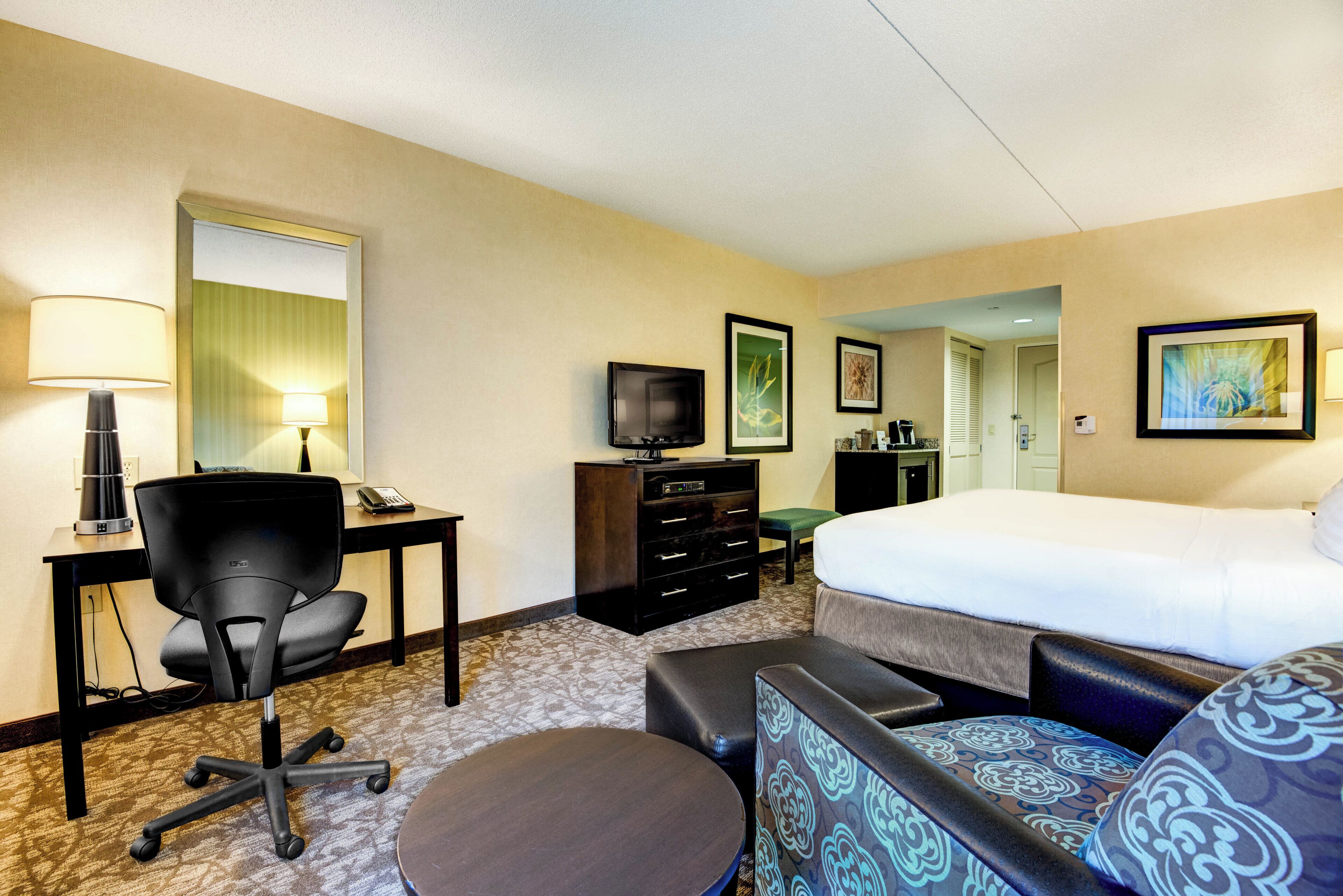 A hotel room with a bed, a desk with a lamp, a chair, a dresser, a television, and a door leading to another room.