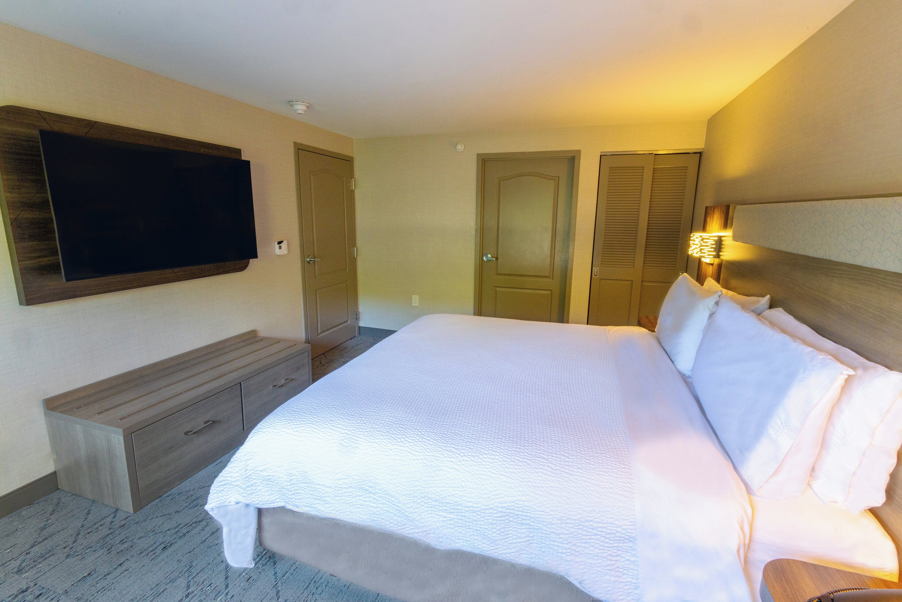 A hotel room with a large bed, a flat-screen TV mounted on the wall, a wooden bench, and a bedside table with a lamp.