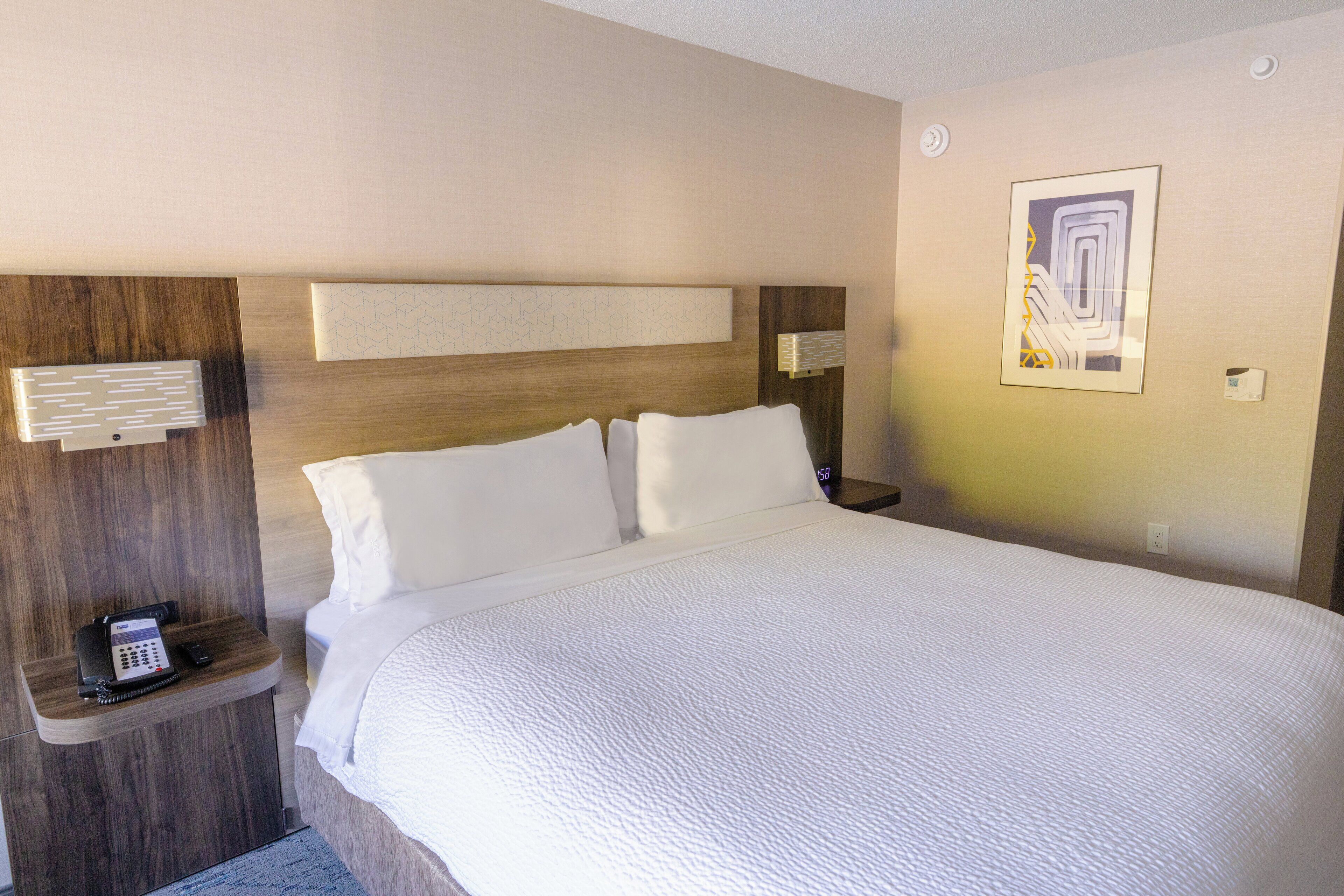 A hotel room with a large bed, a wooden headboard, a bedside table with a phone, and a wall-mounted light.