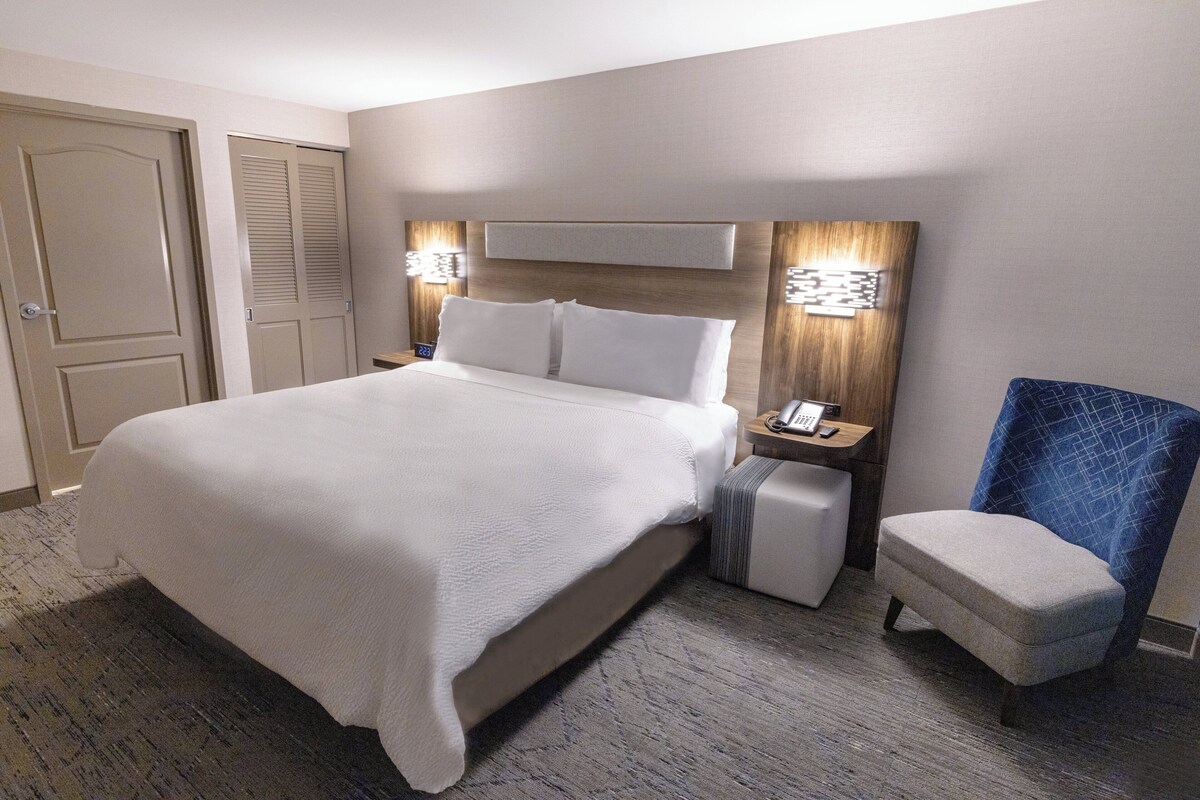 Quarto de hotel com uma cama grande, mesa de cabeceira com telefone, uma cadeira e uma porta.