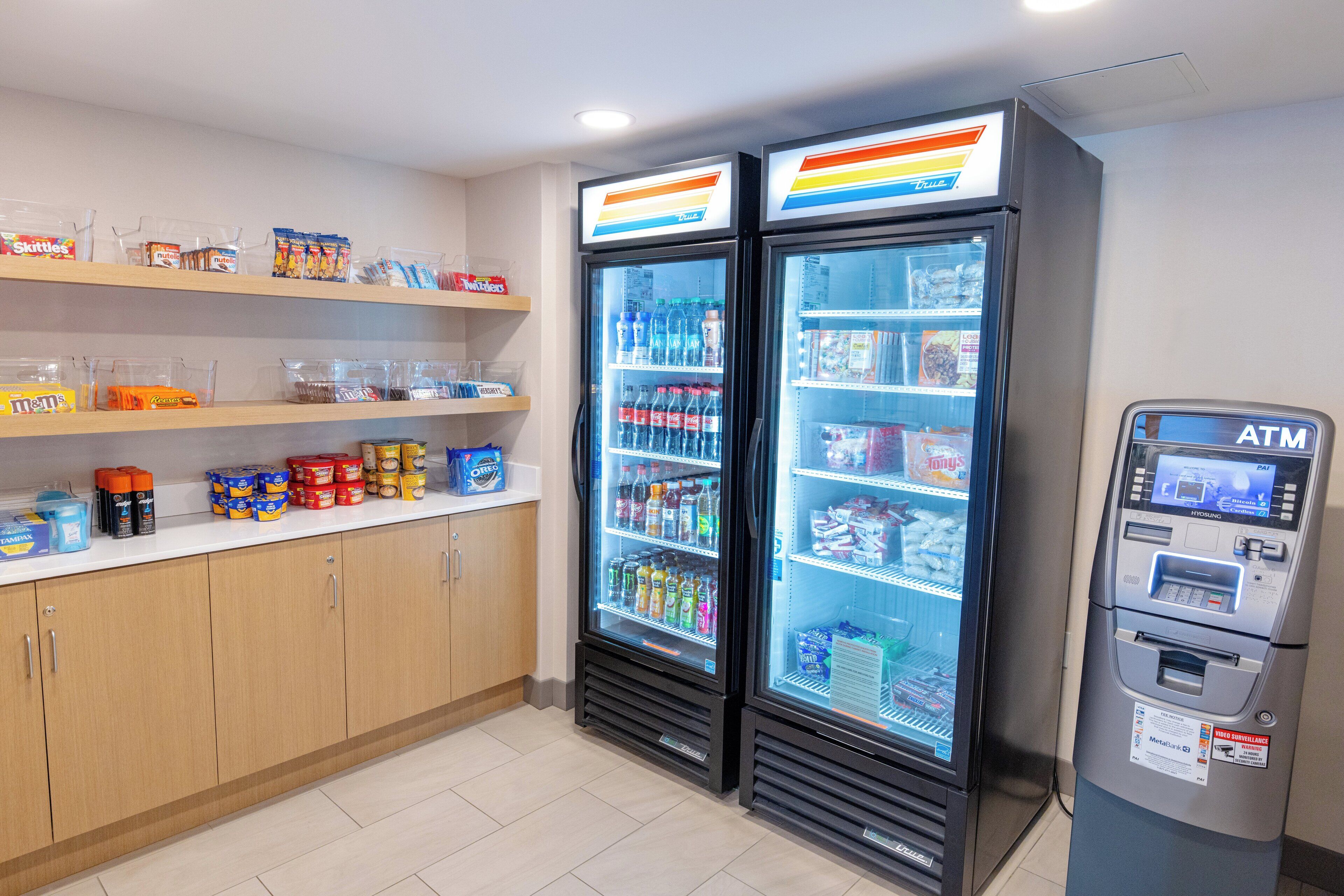 A small convenience store with two refrigerated beverage dispensers, shelves stocked with snacks and drinks, and an ATM machine.