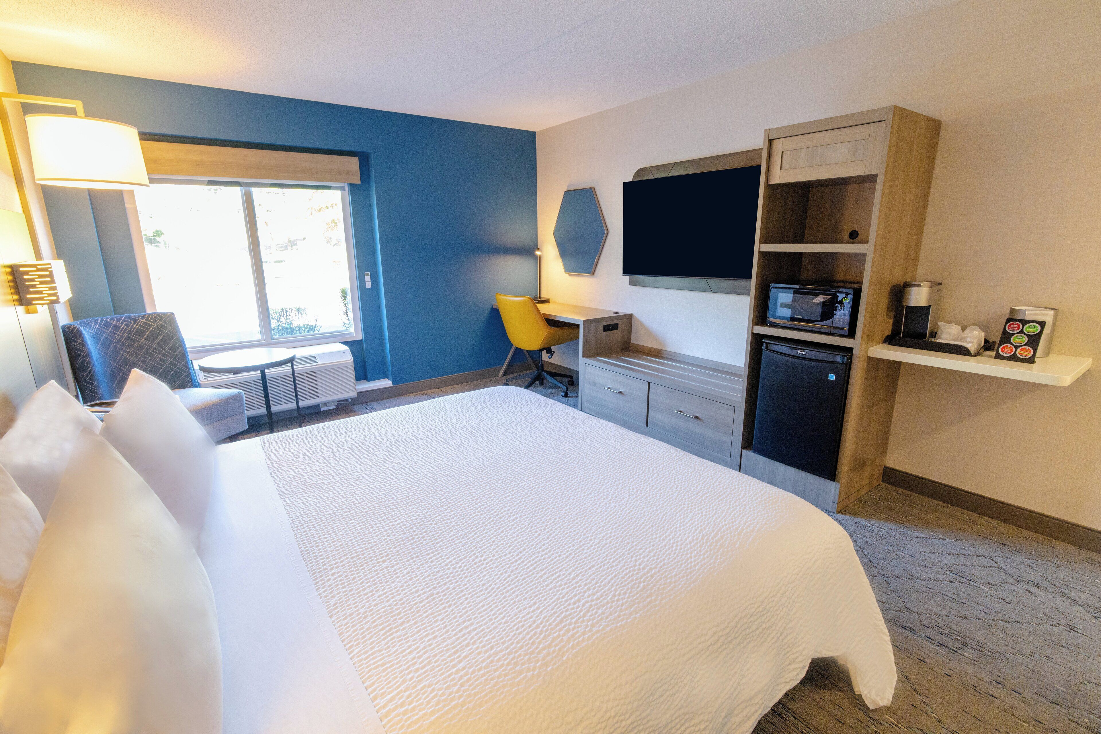 A hotel room with a large bed, a desk with a chair, a flat-screen TV, a microwave, a coffee maker, and a window with blinds.
