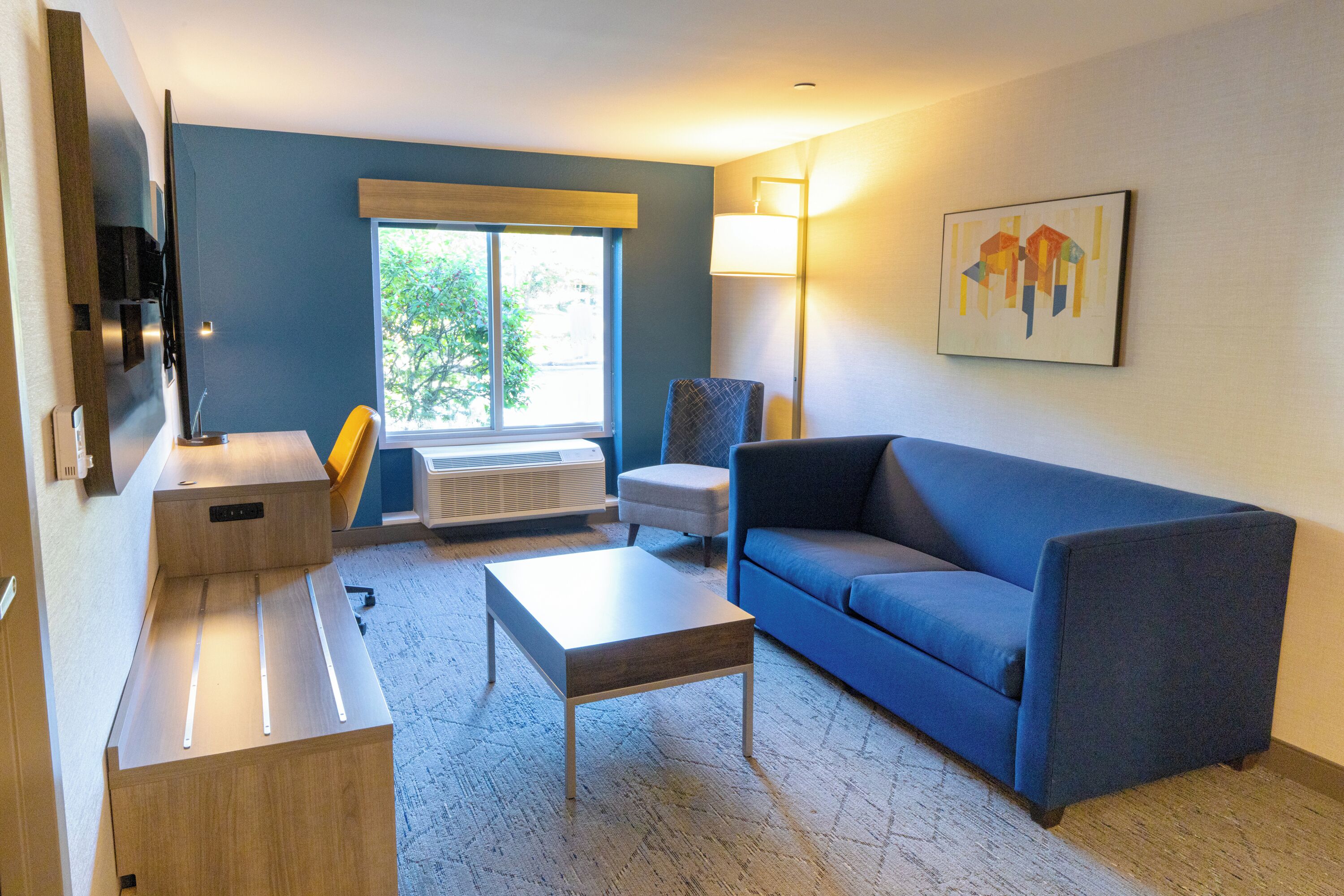 Un salon moderne avec un canapé bleu, une table basse en bois, un petit fauteuil, un téléviseur à écran plat et une fenêtre donnant sur des arbres.