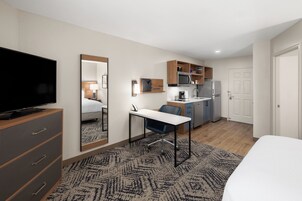 Una habitación de hotel con cama, escritorio con silla, televisión, microondas, refrigerador y una pequeña kitchenette.