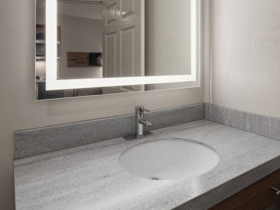 Un baño con encimera de granito, un lavabo blanco, un grifo moderno y un espejo con luz.