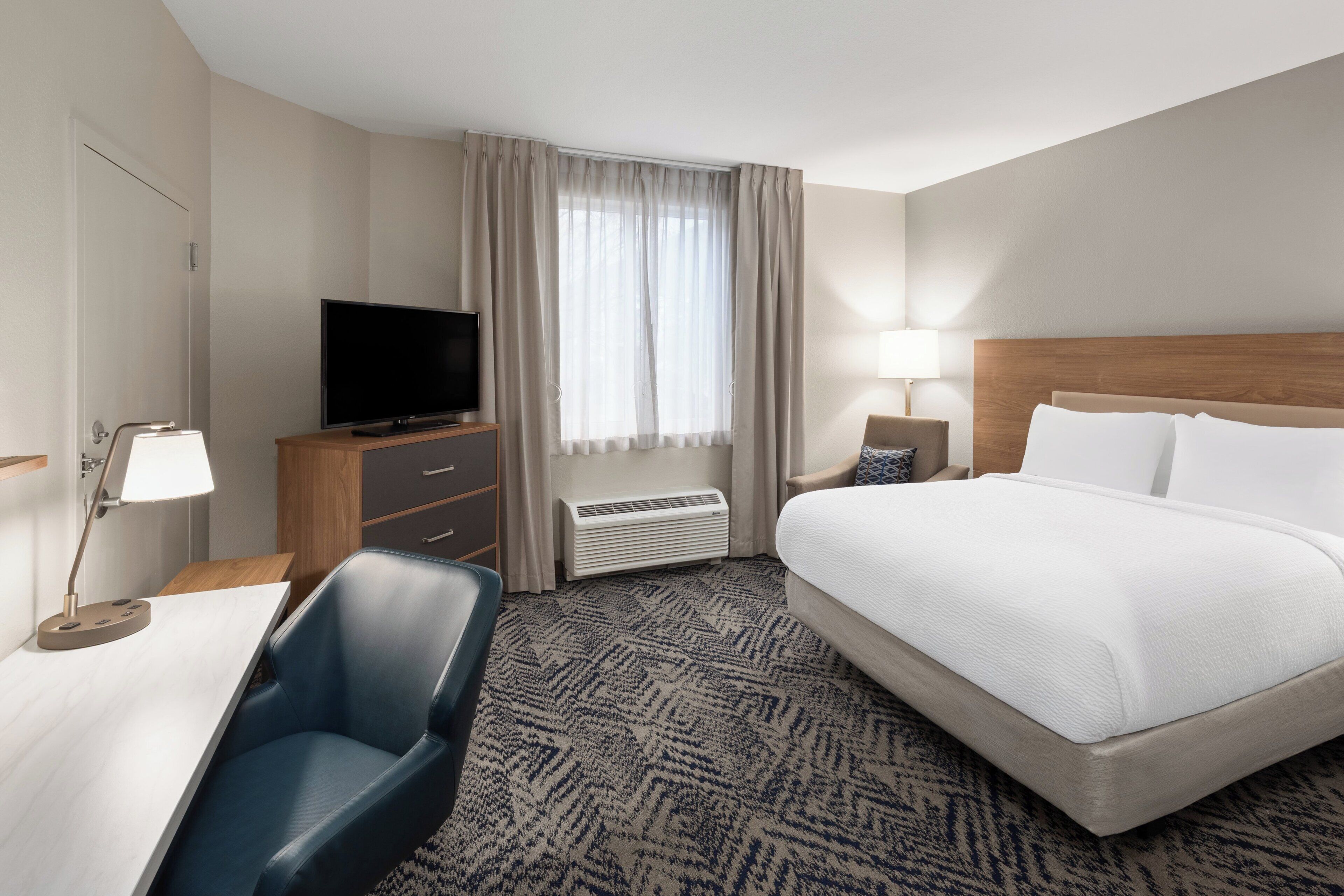 A hotel room with a bed, a desk with a chair, a television, a lamp, and a window with curtains.