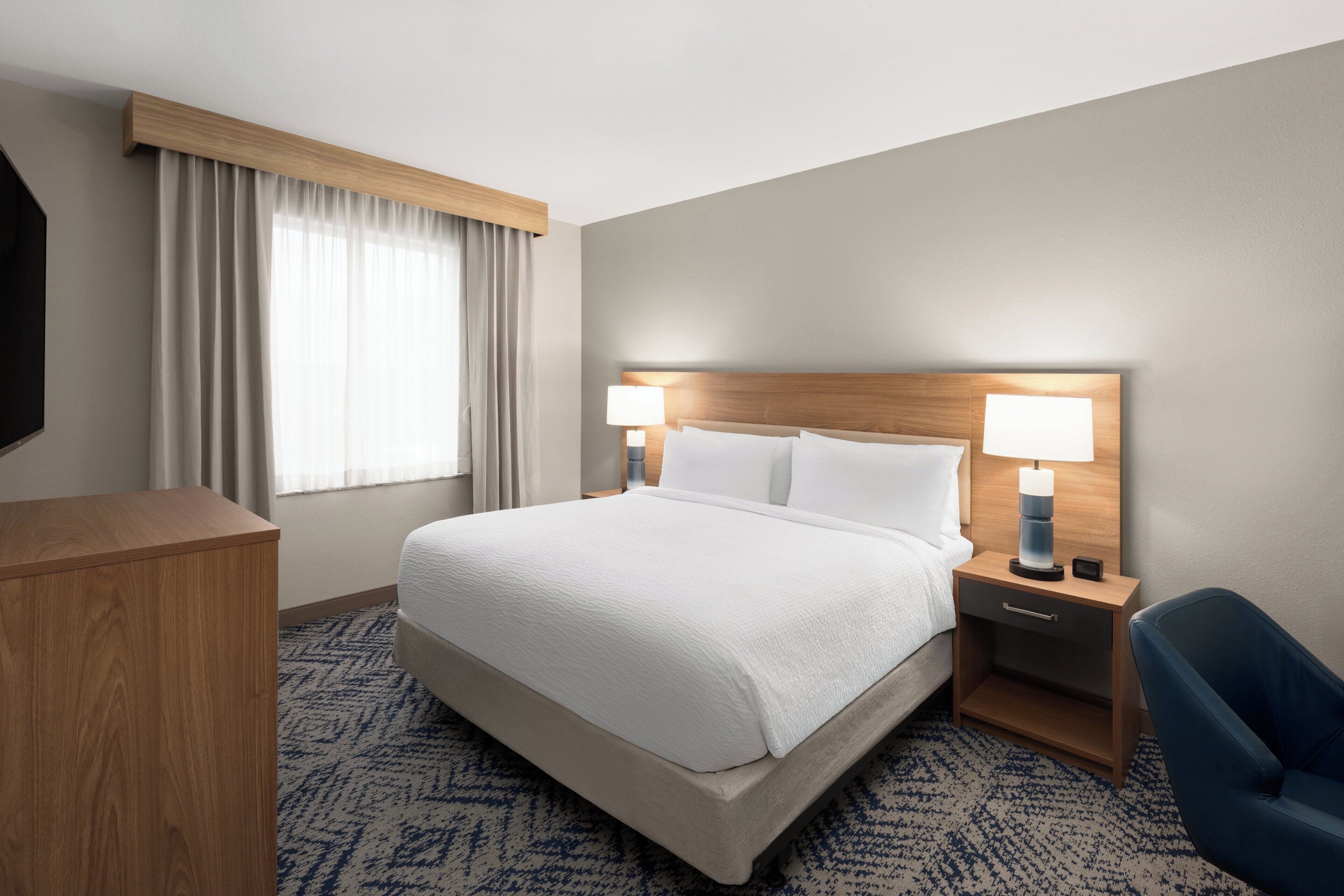 Ein Hotelzimmer mit einem großen Bett, zwei Nachttischen inklusive Lampen, einem Holz-Kopfteil, einem Schreibtisch und einem Stuhl.