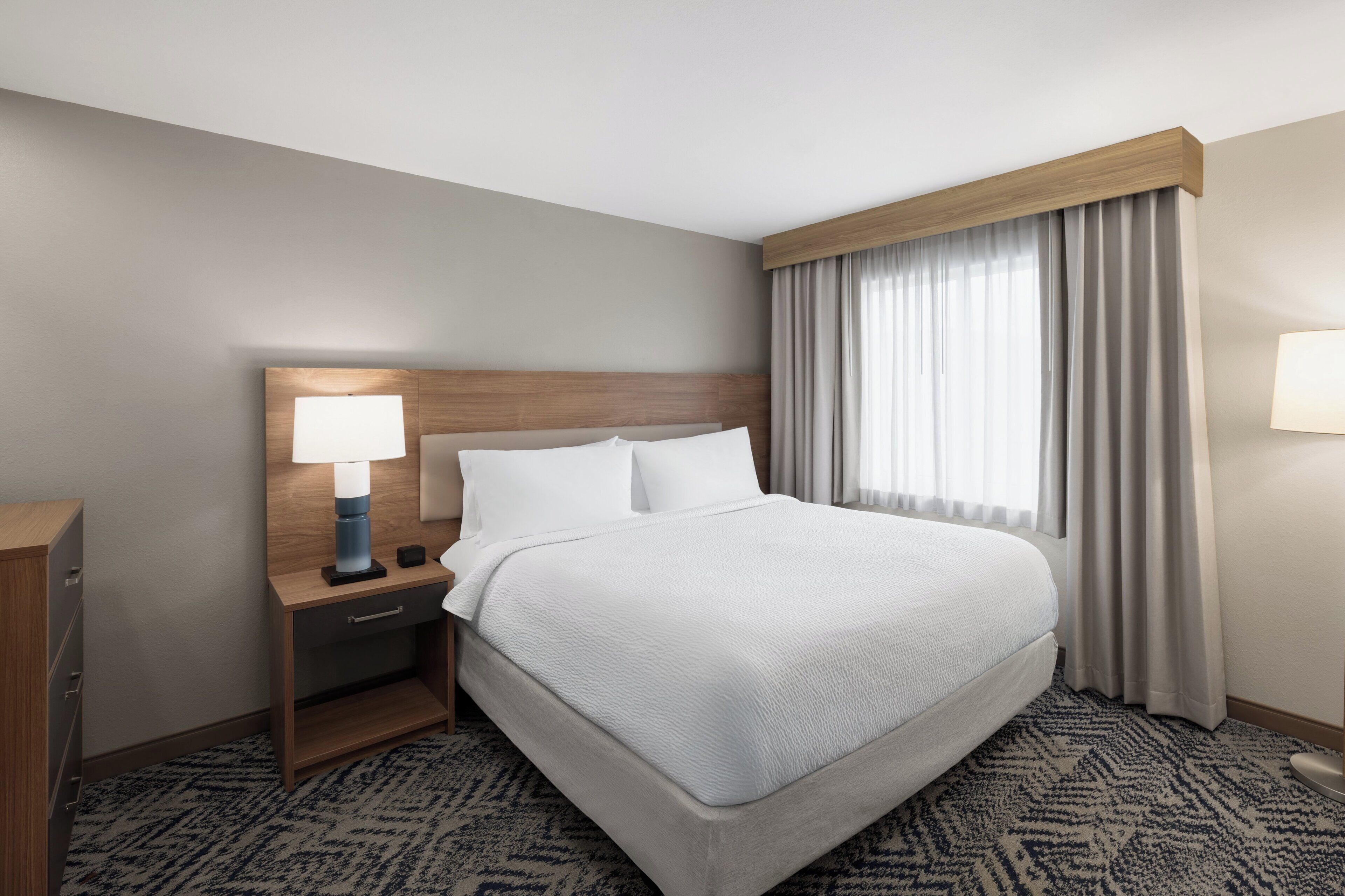 A hotel room with a bed, a nightstand with a lamp, a wooden headboard, and a window with curtains.