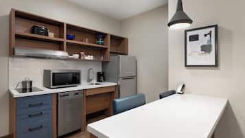 Una cocina moderna con microondas, cafetera y refrigerador. Hay repisas de madera sobre la encimera y una sola silla.